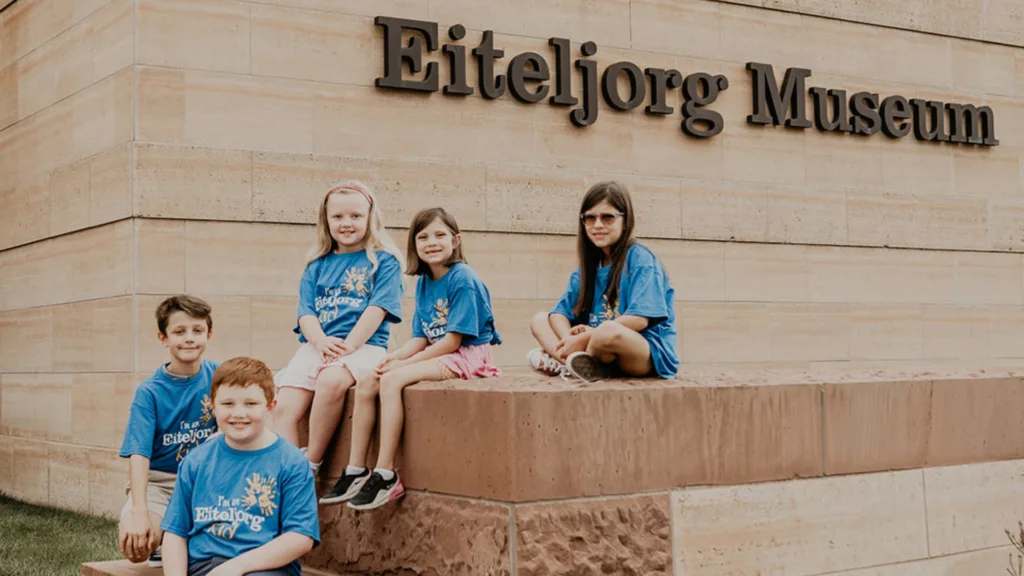 A group of five kids sits on a short wall in front of the Eiteljorg Museum sign.